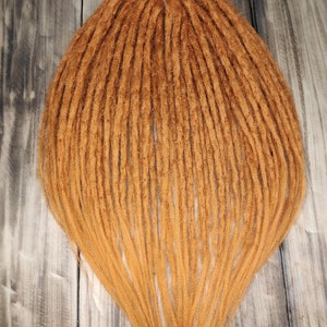 May include: A set of long, brown synthetic dreadlocks. The dreadlocks are arranged in a fan shape and are lying flat on a wooden surface.