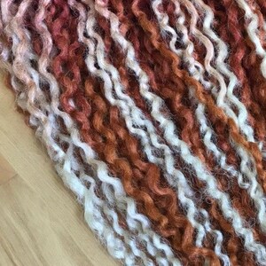 May include: Close-up of a bundle of curly yarn in shades of white and orange-red. The yarn is arranged in rows, creating a textured and colorful pattern.