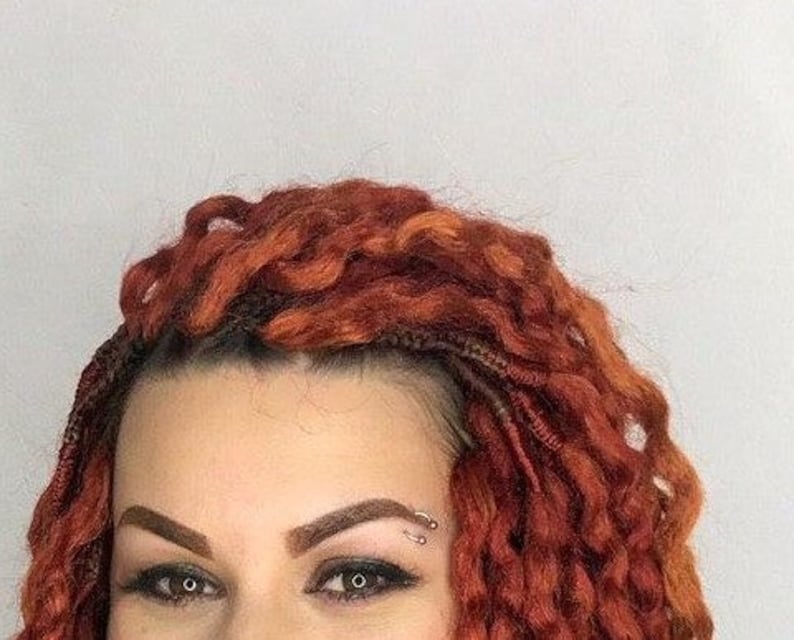 May include: A woman with long, curly, auburn hair. She has a small, silver nose ring and a small, silver ring in her eyebrow.