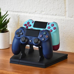 May include: A dark blue wireless game controller positioned in front of a light blue wireless game controller with magenta buttons. Both controllers are resting on a black plastic stand that is mounted on top of a black rectangular gaming console.
