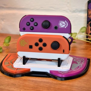 Puede incluir: Dos mandos Joy-Con para Nintendo Switch, uno naranja y blanco y otro morado y blanco, están apilados en un soporte negro y blanco con una base morada y naranja. El soporte tiene un diseño de un escudo con una rueda en su interior.