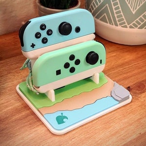 May include: A light blue and green Nintendo Switch Joy-Con controller stand with a white base. The stand is shaped like a beach scene with a green grassy area, a brown sandy beach, and a blue ocean. The stand has a gray rock on the right side and a green leaf logo on the bottom.