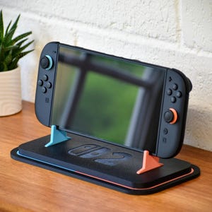 Op de afbeelding: Een zwarte Nintendo Switch-console staat op een zwarte standaard met blauwe en oranje accenten. De standaard heeft een sleuf voor de console en twee steunstukken. Links staat een kleine potplant.