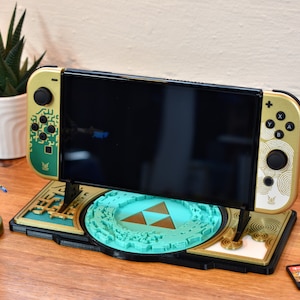 Może przedstawiać: Złota i zielona konsola Nintendo Switch z niestandardowym wzorem inspirowanym grą Zelda. Konsola stoi na stojaku z wzorem Triforce. Obok konsoli znajdują się dwie karty z grami.