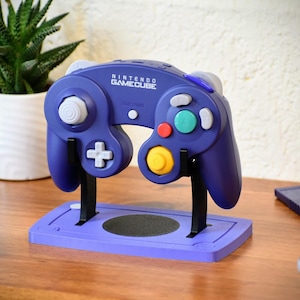 May include: A purple Nintendo GameCube controller on a matching stand. The controller has white, teal, red, and yellow buttons. The stand has two black support arms and a circular base. The words "NINTENDO GAMECUBE" are printed on the controller.