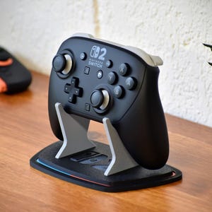May include: Black Nintendo Switch Pro controller on a gray and black stand. The controller has buttons and joysticks. The stand is on a wooden surface. Other controllers are in the background.