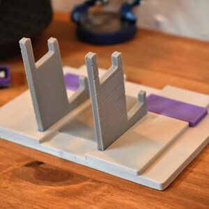 May include: A white plastic stand with two gray slots for holding video game cartridges. The stand has a purple base.