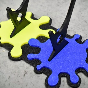 May include: Two black acrylic paintbrush holders stand on a black and yellow paint splatter base. The base is made of foam and has a blue paint splatter on top of the yellow splatter.