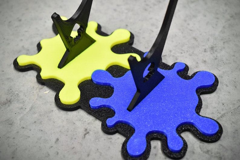 May include: Two black acrylic paintbrush holders stand on a black and yellow paint splatter base. The base is made of foam and has a blue paint splatter on top of the yellow splatter.