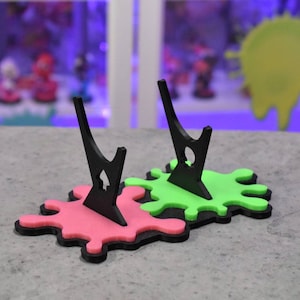 May include: Two black acrylic display stands with a green and pink paint splatter base. The stands are designed to hold figures or other collectibles.
