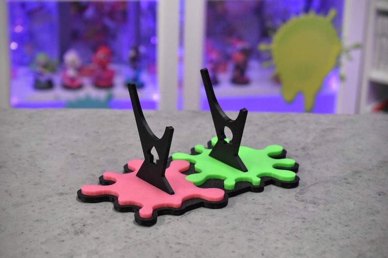 May include: Two black acrylic display stands with a green and pink paint splatter base. The stands are designed to hold figures or other collectibles.