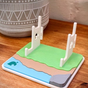May include: White plastic stand with a green, brown, and blue base, resembling a beach scene. The stand has two white pillars with a cutout of a house on the front. The base has a leaf design in the lower left corner.