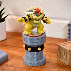 Op de afbeelding: Een gele Bowser-figuur staat op een grijze toren met zwarte stekels. De figuur heeft een rode en groene schild. Een houten kist en een grijs Thwomp-blok zijn ook zichtbaar, in een videogame-thema.