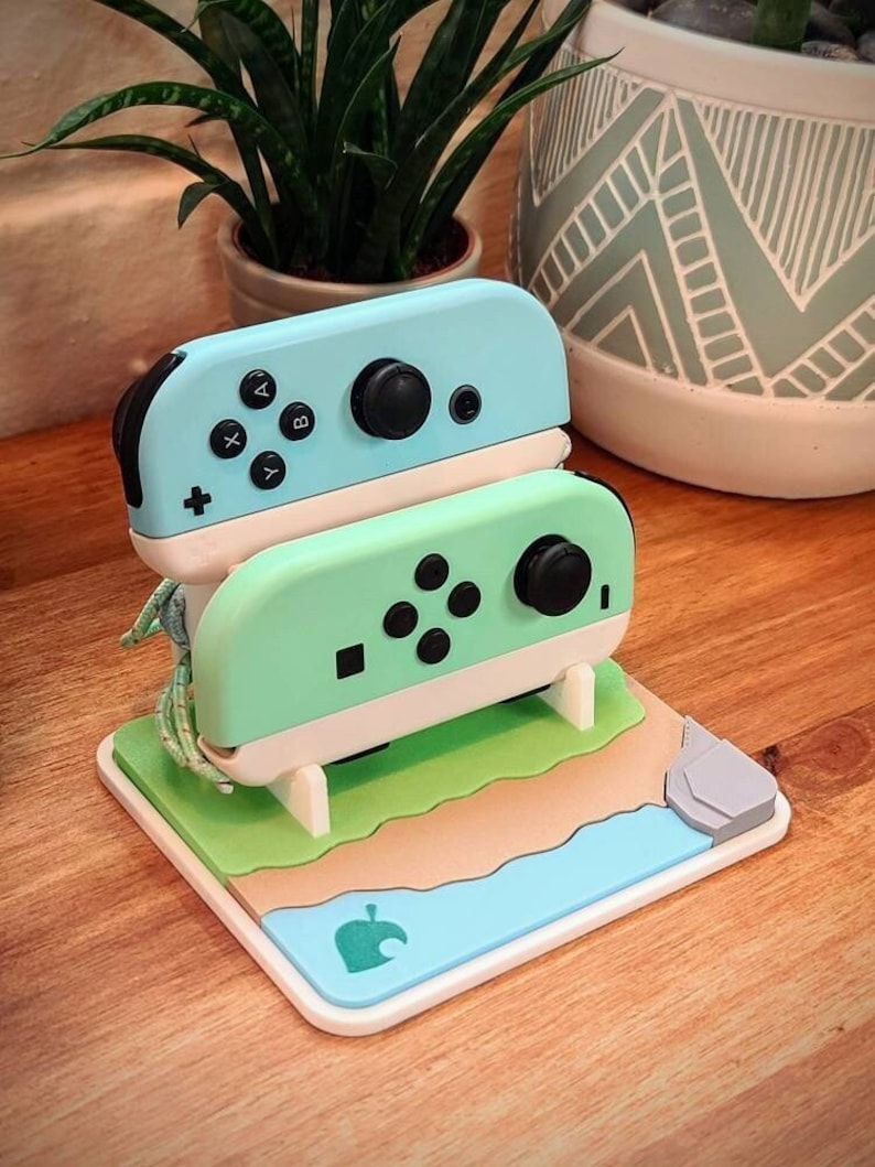 May include: A light blue and green Nintendo Switch Joy-Con controller stand with a white base. The stand is shaped like a beach scene with a green grassy area, a brown sandy beach, and a blue ocean. The stand has a gray rock on the right side and a green leaf logo on the bottom.