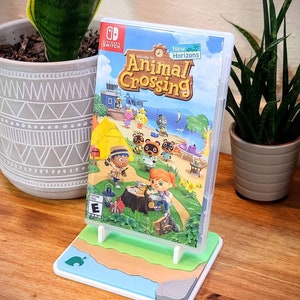 May include: A white and blue video game stand with a green, brown, and blue beach scene. The stand is holding a Nintendo Switch game case for Animal Crossing: New Horizons. The game case features a colorful illustration of characters from the game.
