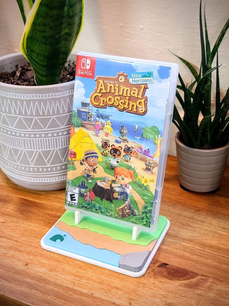 May include: A white and blue video game stand with a green, brown, and blue beach scene. The stand is holding a Nintendo Switch game case for Animal Crossing: New Horizons. The game case features a colorful illustration of characters from the game.