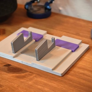 May include: A white plastic stand with two gray slots for holding video game cartridges. The stand has two purple accents on the top.