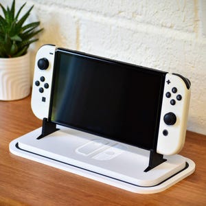Può includere: Una console di videogiochi bianca e nera è esposta su un supporto bianco con supporti neri. La console ha uno schermo nero e controller bianchi. Una piccola pianta in vaso è a sinistra su una superficie di legno.