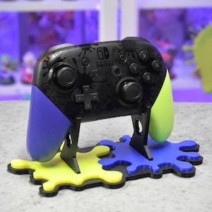 May include: A black Nintendo Switch controller with blue and green paint splatters on the sides, sitting on a black stand with yellow and blue paint splatter bases. The controller has the Nintendo Switch logo on the front.