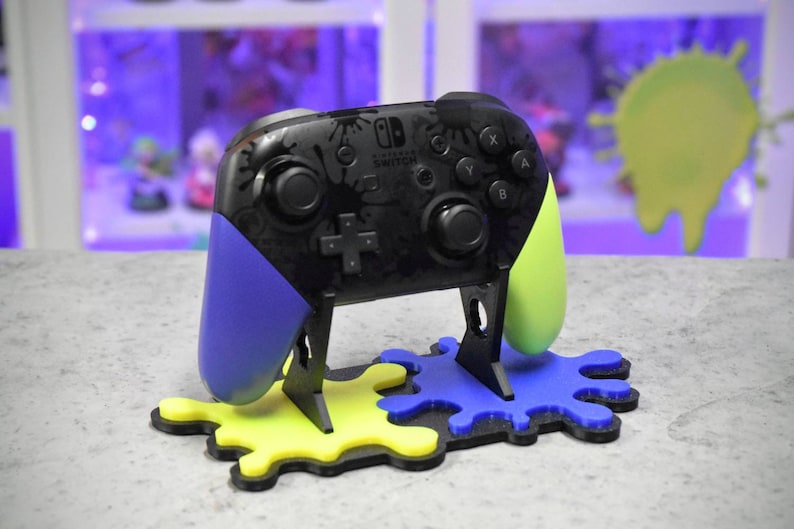 May include: A black Nintendo Switch controller with blue and green paint splatters on the sides, sitting on a black stand with yellow and blue paint splatter bases. The controller has the Nintendo Switch logo on the front.