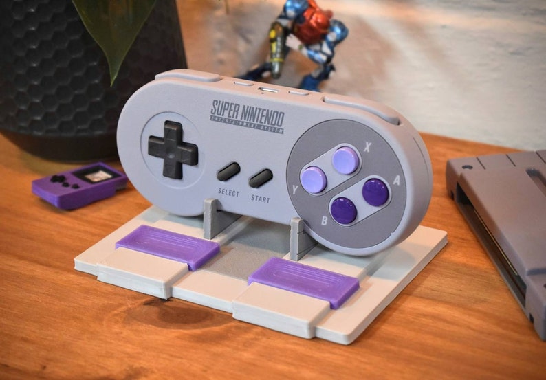 May include: A gray Super Nintendo Entertainment System controller on a white stand with two purple game cartridges. The controller has a black directional pad, four purple buttons, and the text "SUPER NINTENDO" printed on the top.