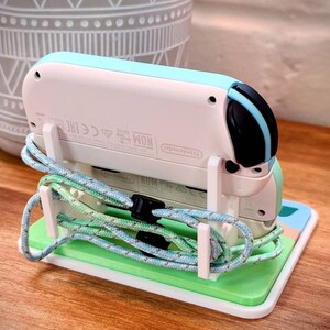 May include: A white and light blue Nintendo Switch console with a black and blue controller, sitting on a green and white stand. The console is connected to a blue and green cord.
