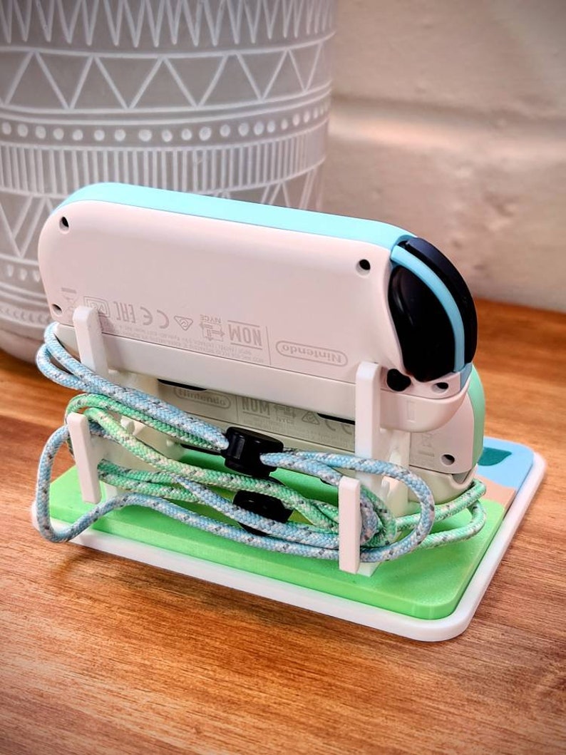 May include: A white and light blue Nintendo Switch console with a black and blue controller, sitting on a green and white stand. The console is connected to a blue and green cord.