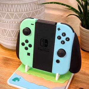 May include: A light blue and green Nintendo Switch console with black Joy-Cons is displayed on a white stand with a green, brown, and blue design. The stand features a stylized leaf icon.
