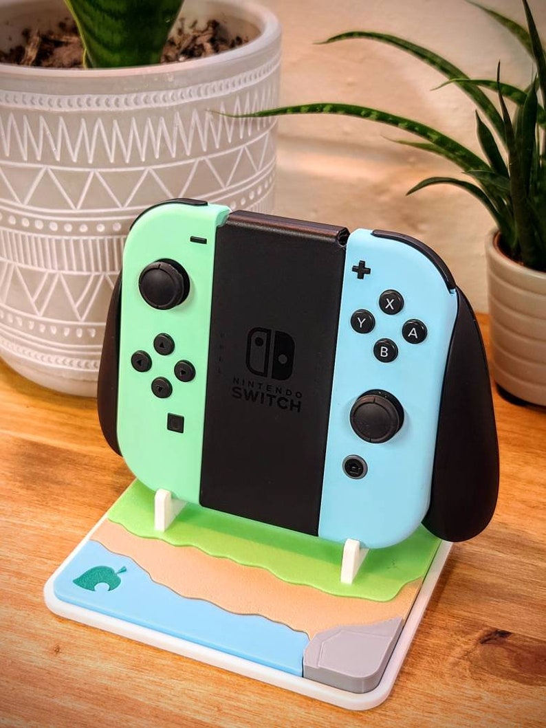 May include: A light blue and green Nintendo Switch console with black Joy-Cons is displayed on a white stand with a green, brown, and blue design. The stand features a stylized leaf icon.
