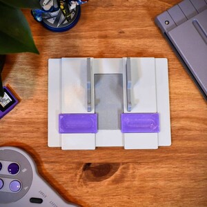 May include: A white plastic stand with two purple slots for holding video game cartridges. The stand is on a wooden surface.