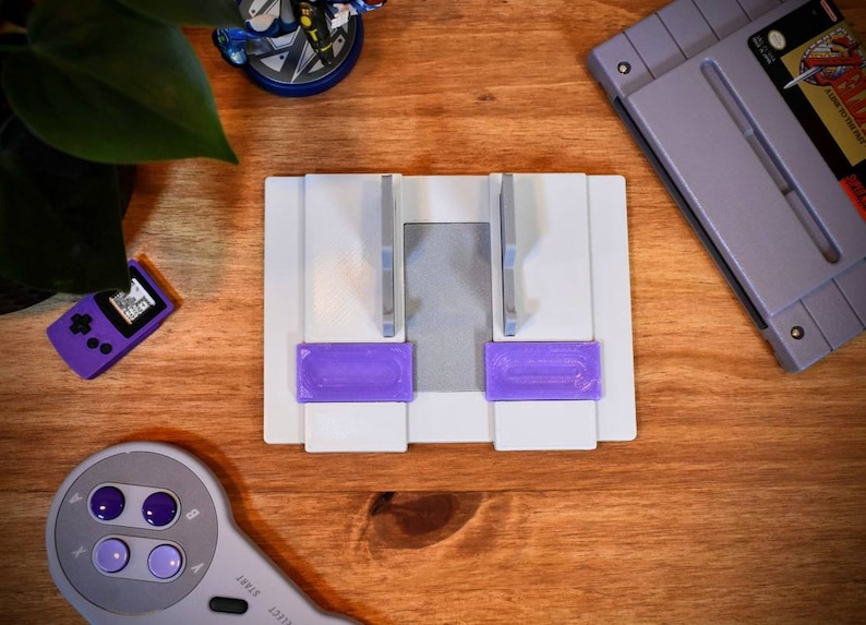 May include: A white plastic stand with two purple slots for holding video game cartridges. The stand is on a wooden surface.