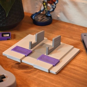 May include: A white and gray Super Nintendo Entertainment System (SNES) game cartridge stand with two slots. The stand has purple accents and is made of plastic. The stand is on a wooden surface.