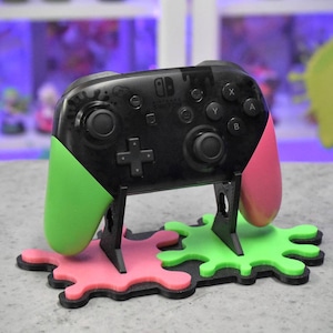 May include: A black Nintendo Switch controller with green and pink Joy-Con grips, sitting on a black stand with green and pink splattered paint designs.
