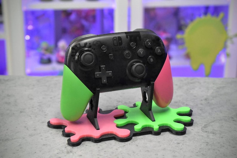 May include: A black Nintendo Switch controller with green and pink Joy-Con grips, sitting on a black stand with green and pink splattered paint designs.