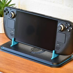 May include: A black handheld gaming console is displayed on a black stand with teal accents. The console has a large screen, buttons, and joysticks. A small potted plant is visible in the background, resting on a wooden surface.