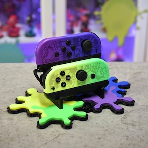 May include: Two purple and green Nintendo Switch Joy-Con controllers with a Splatoon 3 design, sitting on a multi-colored splatter paint stand.