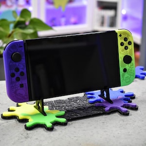 Könnte beinhalten: Eine violette und grüne Nintendo Switch-Konsole mit schwarzem Bildschirm wird auf einem schwarz-weißen 3D-gedruckten Ständer mit bunten Spritzlack-Akzenten präsentiert.