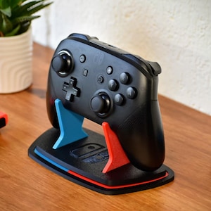 Puede incluir: Mando de videojuegos negro sobre un soporte con detalles azules y rojos. El soporte sostiene el mando en posición vertical. Un mando rojo y otro azul están sobre la mesa. Una pequeña planta en maceta está en el fondo.