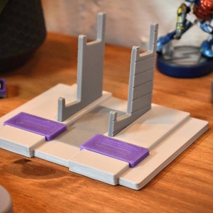 May include: A white plastic stand with two gray vertical supports for holding Super Nintendo game cartridges. The stand has two purple plastic tabs on the front.