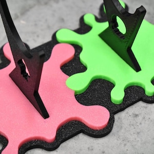 May include: Two black plastic display stands with a green and pink paint splatter design. The stands are made to hold items upright.