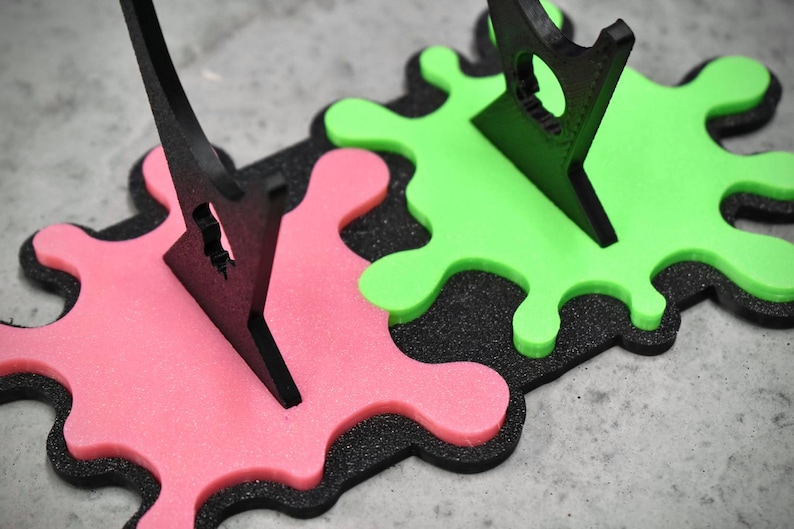 May include: Two black plastic display stands with a green and pink paint splatter design. The stands are made to hold items upright.