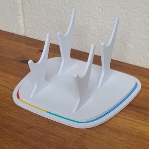 May include: A white plastic stand with four curved supports, designed to hold items. The base is rectangular with rounded corners, featuring a colorful trim of red, yellow, and blue. The stand is on a wooden surface.