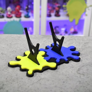 May include: Two black acrylic display stands with a yellow and blue paint splatter design. The stands are made to hold figurines or other collectibles.