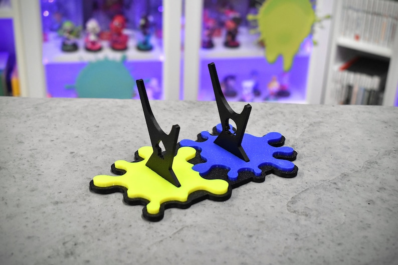 May include: Two black acrylic display stands with a yellow and blue paint splatter design. The stands are made to hold figurines or other collectibles.
