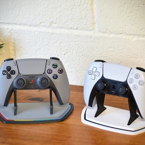 Op de afbeelding: Twee gamecontrollers, één grijs en één wit, tentoongesteld op standaards. De grijze controller heeft een veelkleurige basis, terwijl de witte controller een witte basis heeft. Een kleine potplant staat op de achtergrond.