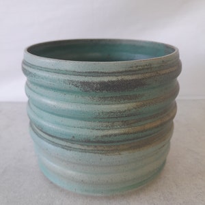 May include: A light blue ceramic planter with a textured, ribbed design. The planter has a subtle, faded look, with a hint of grey.