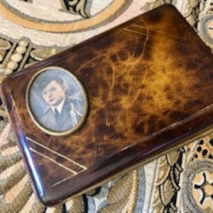 Puede incluir: Un estuche de cigarrillos vintage rectangular de carey marrón con ribete de metal dorado. Un pequeño retrato redondo de un hombre con traje está incrustado en la parte delantera del estuche.