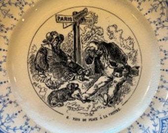 May include: A white ceramic plate with a blue and white floral border. The center of the plate features a black and white illustration of a man and a dog sitting by a signpost that reads "Paris" and "La Chapelle". The text "Le Pain de Place & La Crêpe" is printed below the illustration.