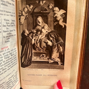 May include: An open book featuring a sepia-toned illustration of the Virgin Mary and baby Jesus, with angels. The text "NOTRE-DAME DU ROSAIRE" is below the image. The book's pages are aged, with gold-gilded edges.
