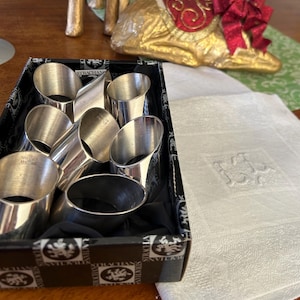 May include: A set of silver napkin rings in a black and white patterned box. The napkin rings are cylindrical with a slight taper. A white linen napkin with an embroidered initial is next to the box.